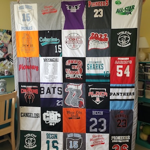 May include: A handmade quilt composed of various sports team t-shirts. The quilt features a patchwork design with different colors, logos, and numbers. The quilt is displayed against a neutral background, showcasing the variety of designs.