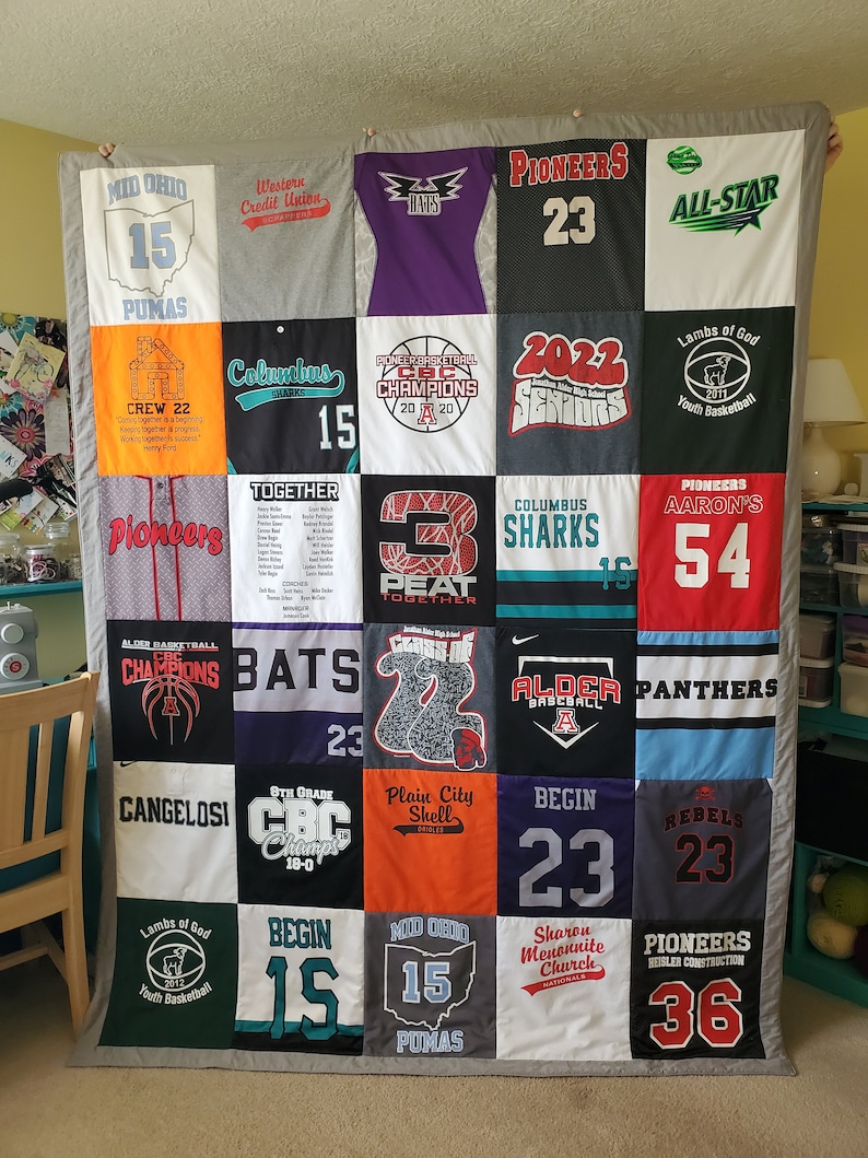 May include: A handmade quilt composed of various sports team t-shirts. The quilt features a patchwork design with different colors, logos, and numbers. The quilt is displayed against a neutral background, showcasing the variety of designs.