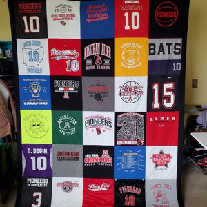 May include: A handmade quilt composed of various t-shirt squares, featuring team names, logos, and numbers. The quilt showcases a variety of colors, including black, red, white, and blue. The text on the shirts includes "Pioneers", "Jonathan Alder", and "Champions".
