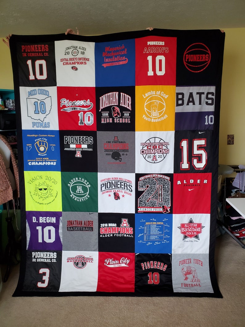 May include: A handmade quilt composed of various t-shirt squares, featuring team names, logos, and numbers. The quilt showcases a variety of colors, including black, red, white, and blue. The text on the shirts includes "Pioneers", "Jonathan Alder", and "Champions".