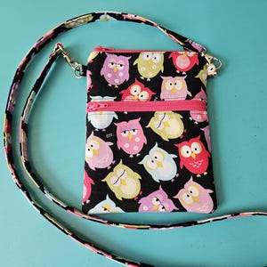 Peut inclure: Un sac bandouli&egrave;re noir avec une fermeture &eacute;clair et une sangle roses. Le sac est orn&eacute; d'illustrations color&eacute;es de hiboux roses, jaunes, bleus et rouges. La sangle pr&eacute;sente un motif avec des accents noirs, roses et violets.
