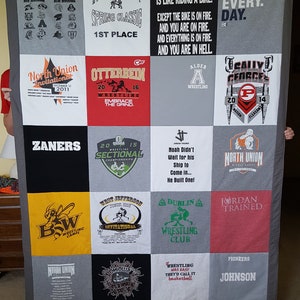 May include: A gray, black, and white quilt made from various t-shirts. The quilt features squares with text and graphics related to wrestling, including team names, event titles, and motivational quotes. The quilt is held up by two people.