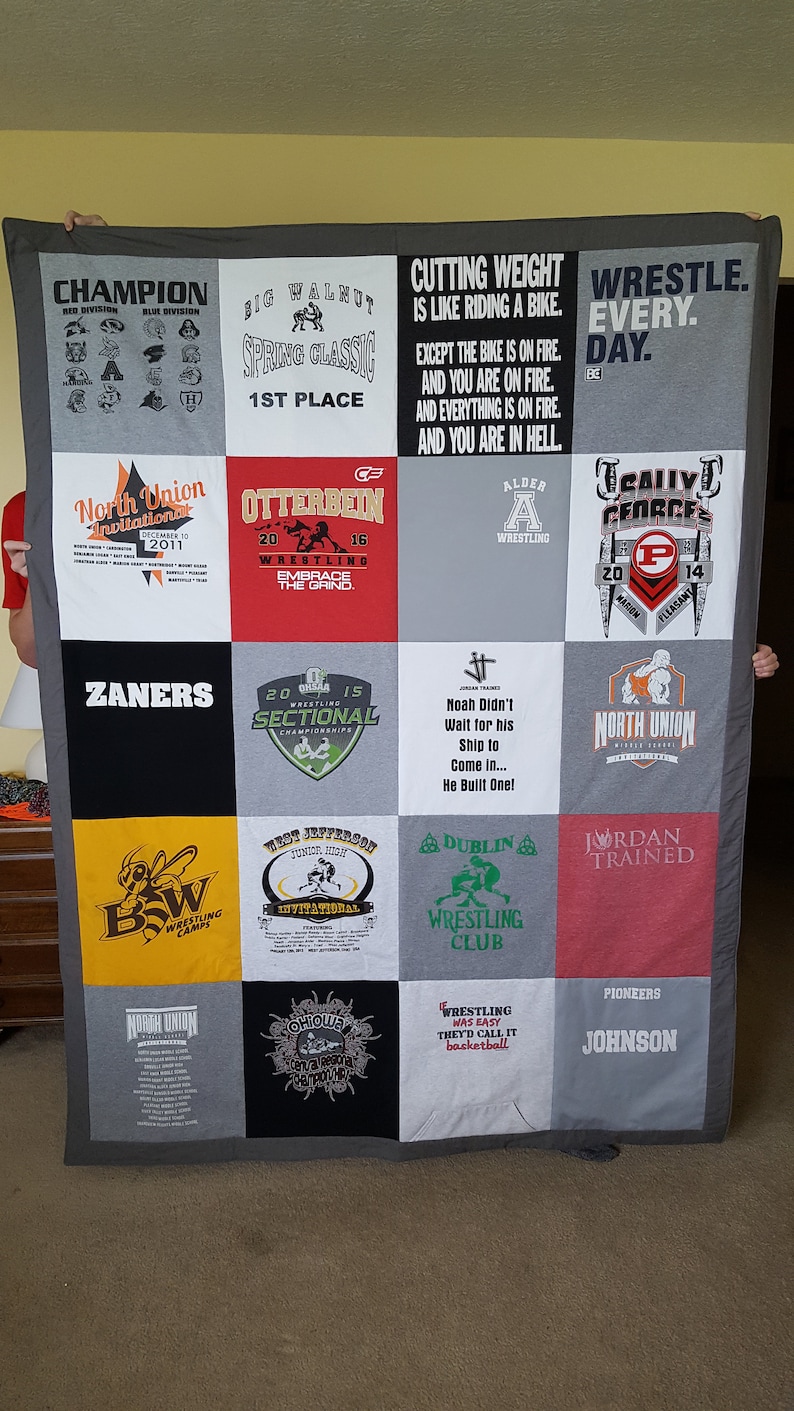 May include: A gray, black, and white quilt made from various t-shirts. The quilt features squares with text and graphics related to wrestling, including team names, event titles, and motivational quotes. The quilt is held up by two people.