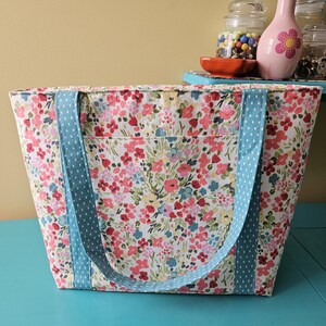 May include: A floral tote bag with light blue polka dot handles. The bag has a colourful pattern of pink, red, and blue flowers on a cream background. The tote is rectangular with a front pocket.