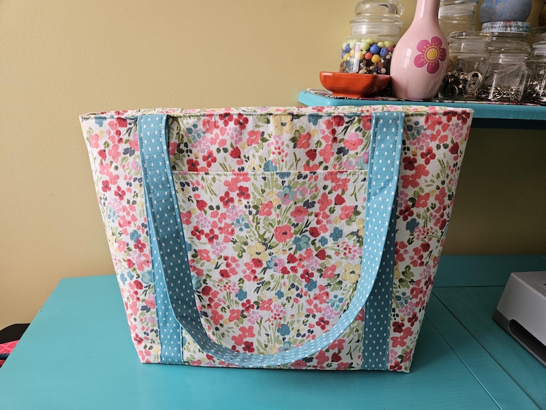 May include: A floral tote bag with light blue polka dot handles. The bag has a colourful pattern of pink, red, and blue flowers on a cream background. The tote is rectangular with a front pocket.