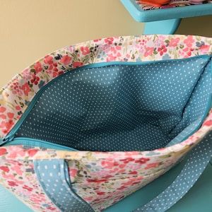 May include: A floral tote bag, viewed from above. The exterior has a white background with pink and red flowers. The interior is teal with white polka dots. A teal zip and straps complete the design.
