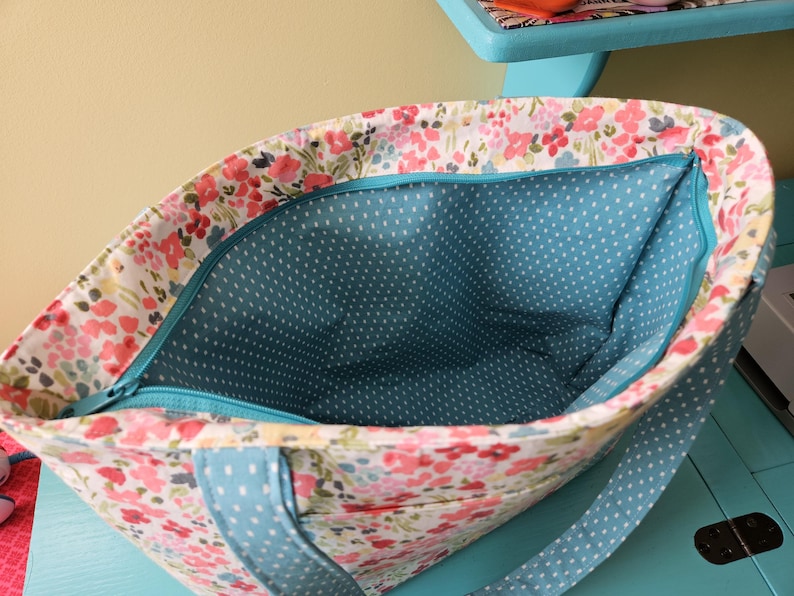 May include: A floral tote bag, viewed from above. The exterior has a white background with pink and red flowers. The interior is teal with white polka dots. A teal zip and straps complete the design.