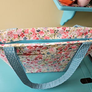 May include: A floral print tote bag with a teal zip and light blue polka dot straps. The bag features a colourful pattern of pink, red, and blue flowers on a white background. The tote has two exterior pockets.