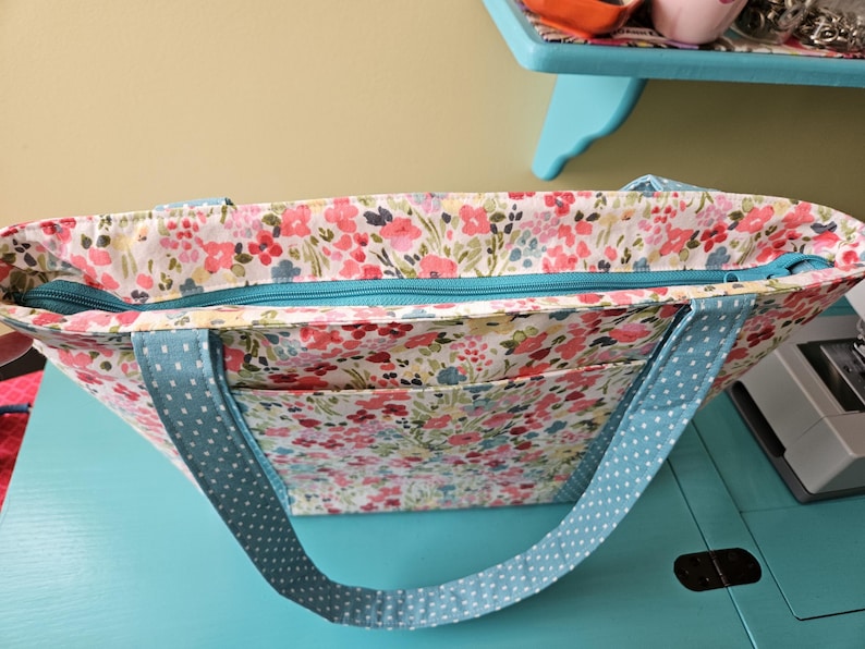 May include: A floral print tote bag with a teal zip and light blue polka dot straps. The bag features a colourful pattern of pink, red, and blue flowers on a white background. The tote has two exterior pockets.