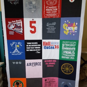 May include: A handmade quilt composed of various t-shirt squares, featuring a white border. The squares display text and graphics, including "French Bulldog's," "Air Force," and "Hall Oates 16." The quilt is a mix of colors.