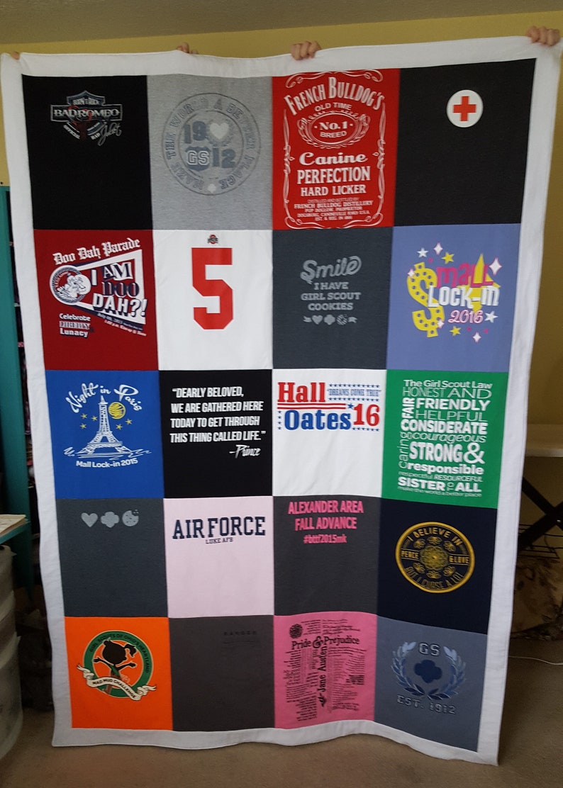 May include: A handmade quilt composed of various t-shirt squares, featuring a white border. The squares display text and graphics, including "French Bulldog's," "Air Force," and "Hall Oates 16." The quilt is a mix of colors.