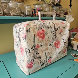 May include: A white sewing machine cover with a Paris themed design. The cover features a pink rose, a black and white Eiffel Tower, and the word "Paris" repeated several times. The cover has a white handle.