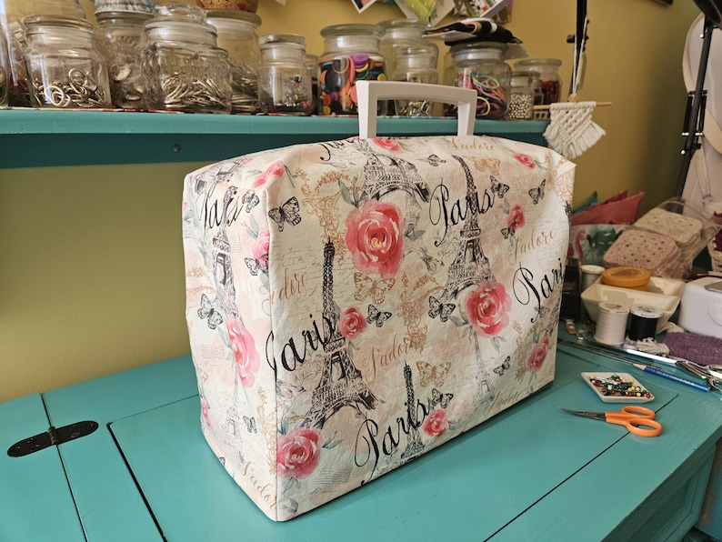 May include: A white sewing machine cover with a Paris themed design. The cover features a pink rose, a black and white Eiffel Tower, and the word "Paris" repeated several times. The cover has a white handle.