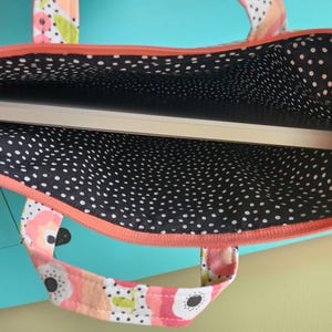 K&ouml;nnte beinhalten: Ein Laptop-Computer in einer Laptop-Tasche mit floralem Muster. Die Tasche hat ein schwarz-wei&szlig;es Polka-Dot-Futter und einen korallenfarbenen Rei&szlig;verschluss. Die Griffe sind mit einem floralen Muster verziert. Die Tasche dient zum Schutz und Transport eines Laptops.