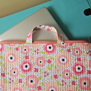 K&ouml;nnte beinhalten: Eine Laptop-H&uuml;lle mit Blumenmuster und Griff. Die H&uuml;lle hat ein Muster aus rosa und pfirsichfarbenen Blumen mit schwarzen Punkten auf wei&szlig;em Hintergrund. Der Rei&szlig;verschluss und der Griff sind in passender Pfirsichfarbe gehalten. Ein silberner Laptop ist teilweise sichtbar.