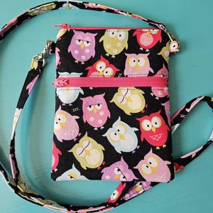 Peut inclure: Un sac bandouli&egrave;re noir avec un motif de hiboux color&eacute;s. Le sac a une fermeture &eacute;clair et une sangle roses, et une sangle assortie avec des motifs de hiboux et de fleurs. Le sac mesure environ 15 cm de large et 20 cm de haut.