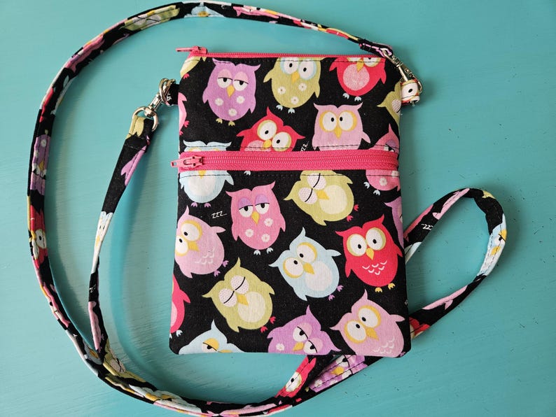 Peut inclure: Un sac bandouli&egrave;re noir avec un motif de hiboux color&eacute;s. Le sac a une fermeture &eacute;clair et une sangle roses, et une sangle assortie avec des motifs de hiboux et de fleurs. Le sac mesure environ 15 cm de large et 20 cm de haut.