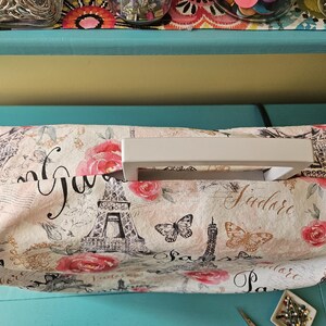 May include: A white sewing machine cover with a Parisian-themed print featuring the Eiffel Tower, roses, butterflies, and the text "J'adore Paris". The cover has a white handle.