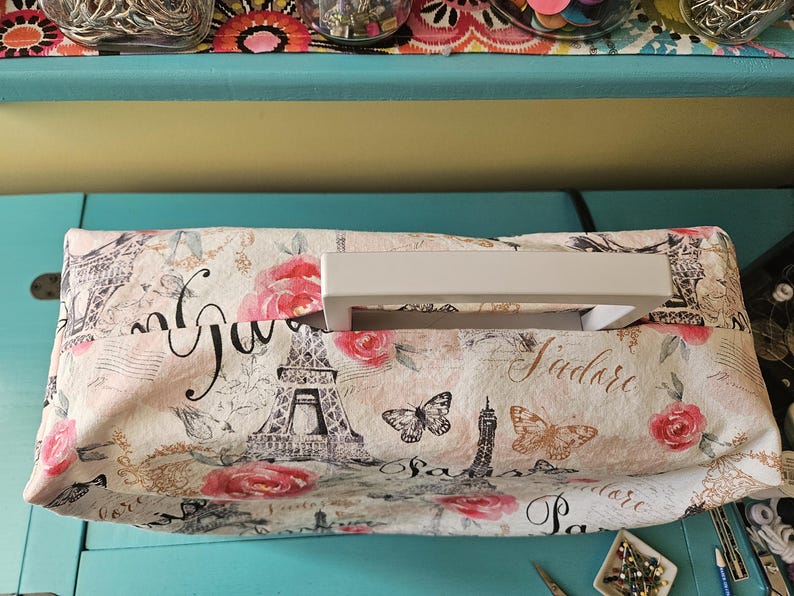 May include: A white sewing machine cover with a Parisian-themed print featuring the Eiffel Tower, roses, butterflies, and the text "J'adore Paris". The cover has a white handle.