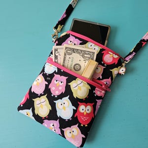 Peut inclure: Sac bandouli&egrave;re noir avec un motif de hiboux color&eacute;s, avec des fermetures &eacute;clair et une sangle roses. Le sac est ouvert, r&eacute;v&eacute;lant un smartphone, de l'argent et un petit porte-cartes. Le fond est de couleur turquoise.