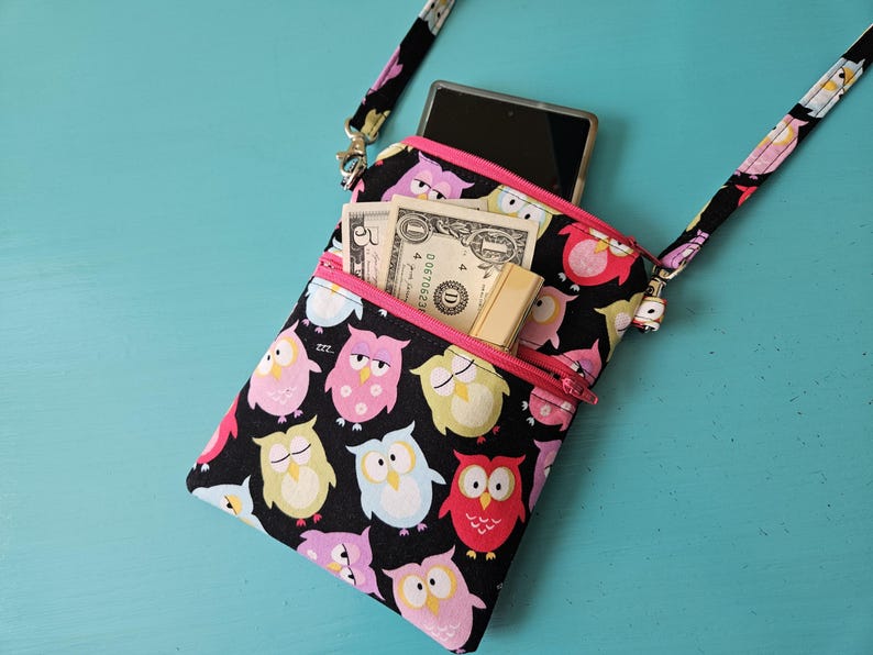 Peut inclure: Sac bandouli&egrave;re noir avec un motif de hiboux color&eacute;s, avec des fermetures &eacute;clair et une sangle roses. Le sac est ouvert, r&eacute;v&eacute;lant un smartphone, de l'argent et un petit porte-cartes. Le fond est de couleur turquoise.