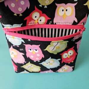 Peut inclure: Un petit sac ouvert avec un fond noir recouvert de hiboux de dessins anim&eacute;s color&eacute;s. Le sac a une fermeture &eacute;clair rose et un int&eacute;rieur &agrave; rayures noires et blanches. Une dragonne est attach&eacute;e sur le c&ocirc;t&eacute;.