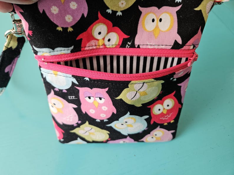 Peut inclure: Un petit sac ouvert avec un fond noir recouvert de hiboux de dessins anim&eacute;s color&eacute;s. Le sac a une fermeture &eacute;clair rose et un int&eacute;rieur &agrave; rayures noires et blanches. Une dragonne est attach&eacute;e sur le c&ocirc;t&eacute;.