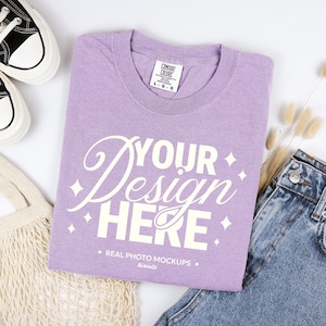 May include: A lavender Comfort Colors t-shirt with the text "YOUR Design HERE" in white. The shirt is laid flat next to a pair of blue jeans, a pair of black and white sneakers, and a beige tote bag. The shirt is a size L-G-G.
