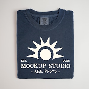 May include: Navy blue t-shirt with a white sunburst design and the text "MOCKUP STUDIO • REAL PHOTO." The shirt is folded neatly and has a tag that says "COMFORT COLORS".