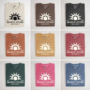 Puede incluir: Nueve camisetas dobladas en varios colores, incluyendo beige, marrón y azul. Cada camiseta presenta un gráfico de sol con el texto "MOCKUP STUDIO - REAL PHOTO" y el año 2026.