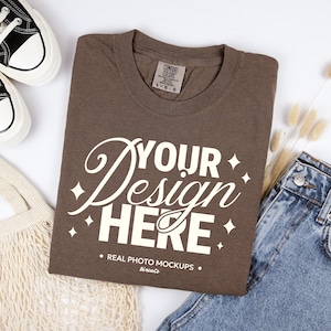 May include: A brown t-shirt with the text "YOUR Design HERE" in a white, stylized font. The shirt is laid flat with a pair of blue jeans, canvas shoes, and a mesh bag. The shirt has a Comfort Colors tag.