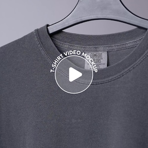 Op de afbeelding: Close-up van een houtskoolgrijs t-shirt dat aan een zwarte hanger hangt. Het shirt heeft een ronde hals en een label met de tekst "COMFORT COLORS". Een witte cirkel met een afspeelknop en de tekst "T-SHIRT VIDEO MOCKUP" is over het shirt geplaatst.