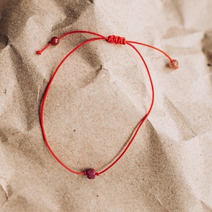 Pode incluir: Uma pulseira de cordão vermelho com uma única conta pequena de pedra preciosa em tom rosa-avermelhado escuro no centro. A pulseira inclui um fecho de nó deslizante ajustável e duas contas menores nas extremidades do cordão.