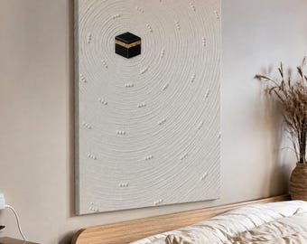 May include: A rectangular wall art piece featuring a textured off-white surface with concentric circular patterns and small raised dots. A small, black cube with a gold horizontal band is positioned off-center on the textured canvas.