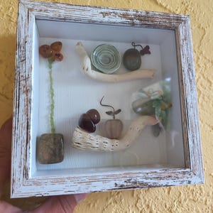 Könnte beinhalten: Ein gerahmter Schattenkasten mit weißem, abgenutztem Finish. Im Inneren sind verschiedene natürliche Elemente angeordnet, darunter eine Blume aus orangefarbenem Glas, ein Schneckenhaus und polierte Steine. Die Anordnung befindet sich vor einem weißen Hintergrund.