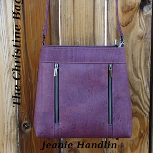 May include: A plum-colored handbag with two front zippered pockets and a matching shoulder strap. The bag is made of a textured material and features the text "The Christine Bag" and "Jeanie Handlin".