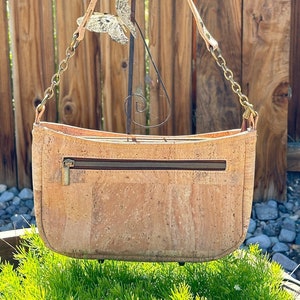 May include: A light brown cork handbag with a gold chain strap and a zipper closure. The bag is hanging from a metal hook.