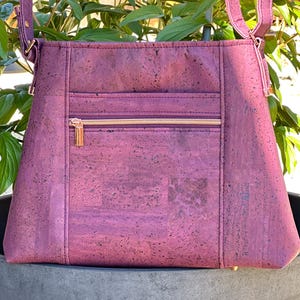 Peut inclure: Un sac à main en liège violet avec une poche zippée et un tirette de fermeture éclair dorée. Le sac a une longue bandoulière et est fait d'un matériau texturé.