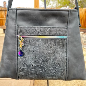 May include: Black leather shoulder bag with a floral embossed design and a rainbow zipper. The bag has a small, round, iridescent charm attached to the zipper.