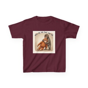 May include: A maroon t-shirt featuring a graphic of two monkeys hugging. The image includes the text "When life hits hard, stay safe." The design is on a square, light-colored background. The shirt is made of a soft material.