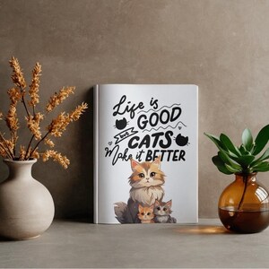 May include: A white book with the text "Life is good but cats make it better" and a cat illustration. The book features a fluffy cat with two kittens. A vase with dried flowers and a vase with a plant are on either side.