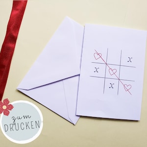 May include: A white card with a tic-tac-toe game drawn with hearts and X's, next to a white envelope. A red ribbon and a graphic with the text "zum DRUCKEN" are also in the image.
