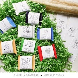 Op de afbeelding: Een assortiment kleurrijke chocoladerepen met Pasen-thema illustraties en tekst, genesteld in groen papier. De wrappers tonen tekeningen van konijntjes, eieren en bloemen. De tekst "DIGITALER DOWNLOAD" staat onderaan.