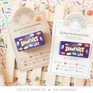 Peut inclure: Deux cartes rectangulaires ornées d'une illustration d'arc-en-ciel et de fleurs, avec le texte "AUF EINEN KUNTERBUNTEN KINDERTAG" et "MIT GANZ VIEL" suivi d'une liste de mots. Chaque carte porte une petite boîte bleue de bonbons Smarties mini.