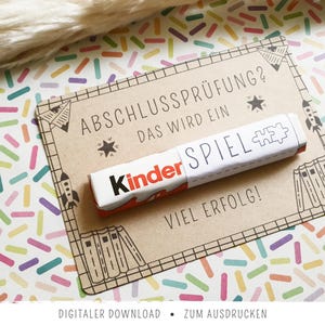 Könnte beinhalten: Ein Kinder-Schokoriegel liegt auf einer braunen Karte mit dem Text "ABSCHLUSSPRÜFUNG? DAS WIRD EIN SPIEL! VIEL ERFOLG!" und dekorativen Zeichnungen. Der Hintergrund ist ein farbenfrohes Muster aus pastellfarbenen Rechtecken.
