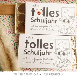 May include: Two rectangular white boxes containing round chocolates, featuring the German text 'Auf ein tolles Schuljahr' and 'und eine aufregende Zeit voller Lernen, Lesen & Lachen', accompanied by line art illustrations of a globe, books, and a chalkboard.