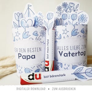 Puede incluir: Dos recipientes de papel blanco con diseños florales y de corazón azules, uno con "FÜR DEN BESTEN Papa" y el otro con "ALLES LIEBE ZUM Vatertag". También es visible una barra de chocolate con el texto "du bist bärenstark" y "bist mein Held". Descarga digital.
