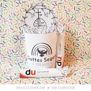 Può includere: Un artigianato di carta bianca con un disegno a tratto nero di una chiesa, una colomba e un arcobaleno. Il testo include "Gottes Segen", "du", "FÜR DICH", "bist wertvoll" e "wirst getragen". Lo sfondo è un motivo colorato di piccoli rettangoli.