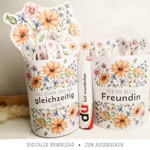 May include: Two cylindrical paper containers decorated with watercolor-style orange and blue wildflowers. The left container features the text "VOLL SCHÖN, DASS WIR gleichzeitig LEBEN" and the right container features "FÜR DIE BESTE Freundin".