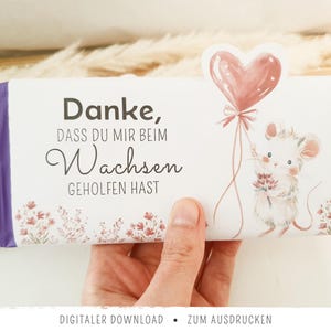 Może przedstawiać: Prostokątne opakowanie na tabliczkę czekolady z białym tłem i fioletowymi końcami. Tekst brzmi Danke, DASS DU MIR BEIM Wachsen GEHOLFEN HAST. Ilustracja przedstawia małą myszkę trzymającą różowy balon w kształcie serca oraz kwiaty.